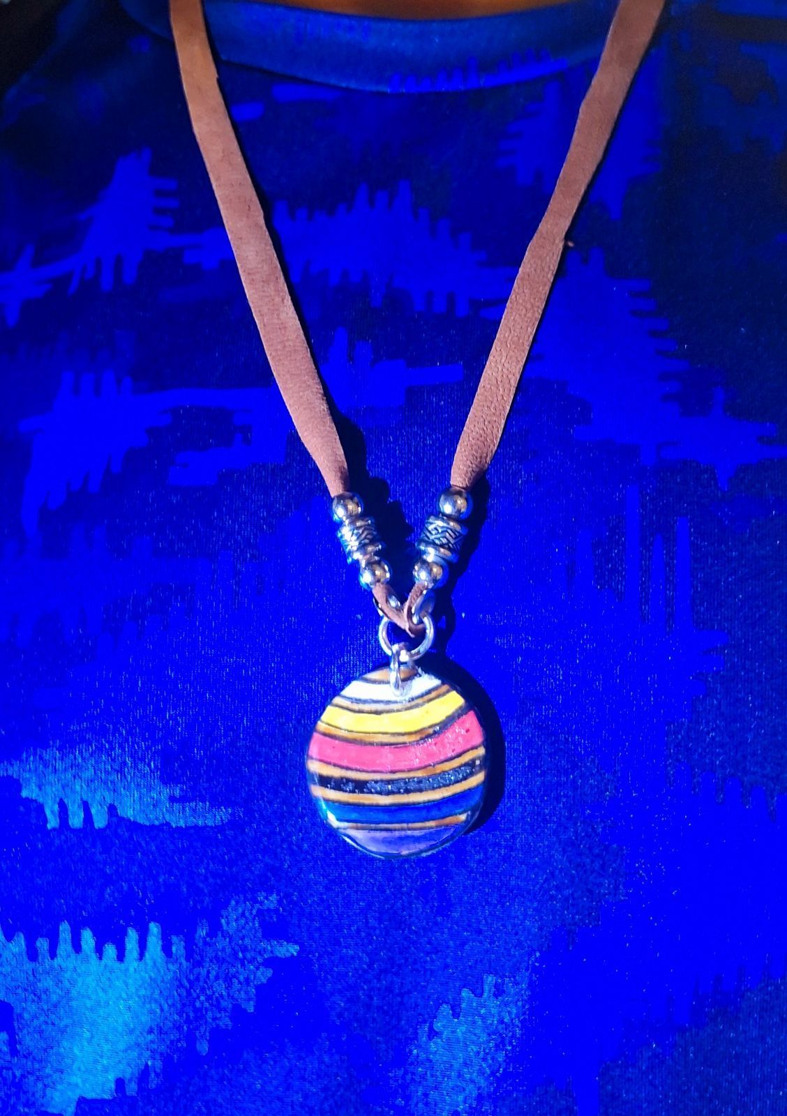 Close-up of a person wearing a necklace with a round pendant. The pendant has colorful stripes, and a brown cord.