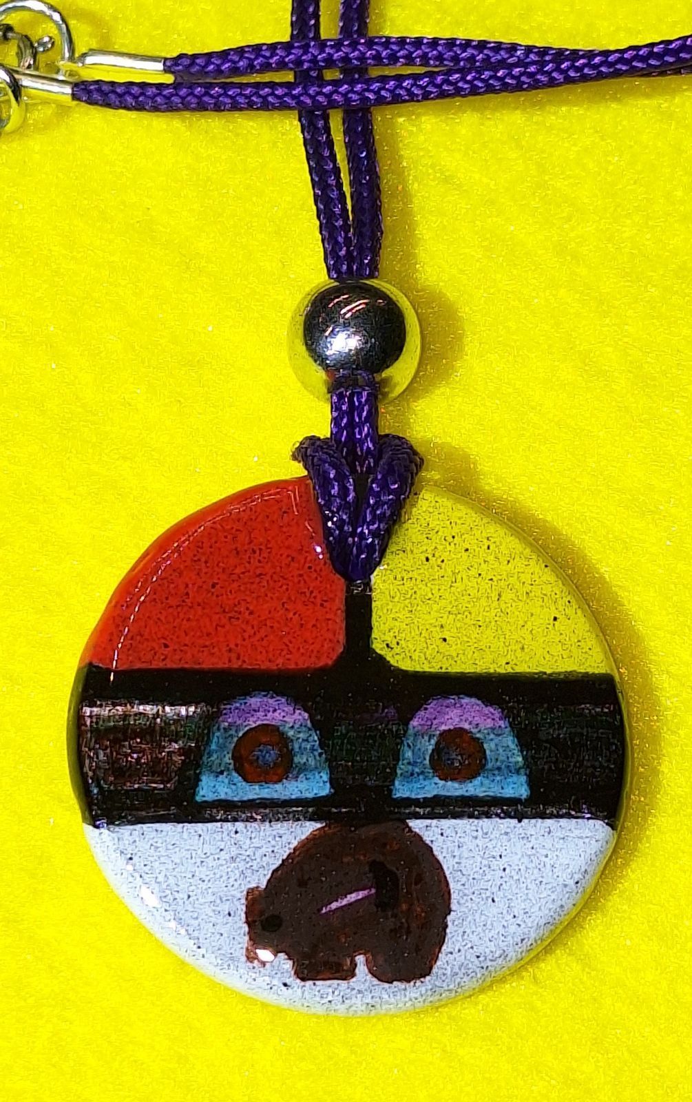Colorful circular pendant with abstract face design; red, yellow, black, and white sections.