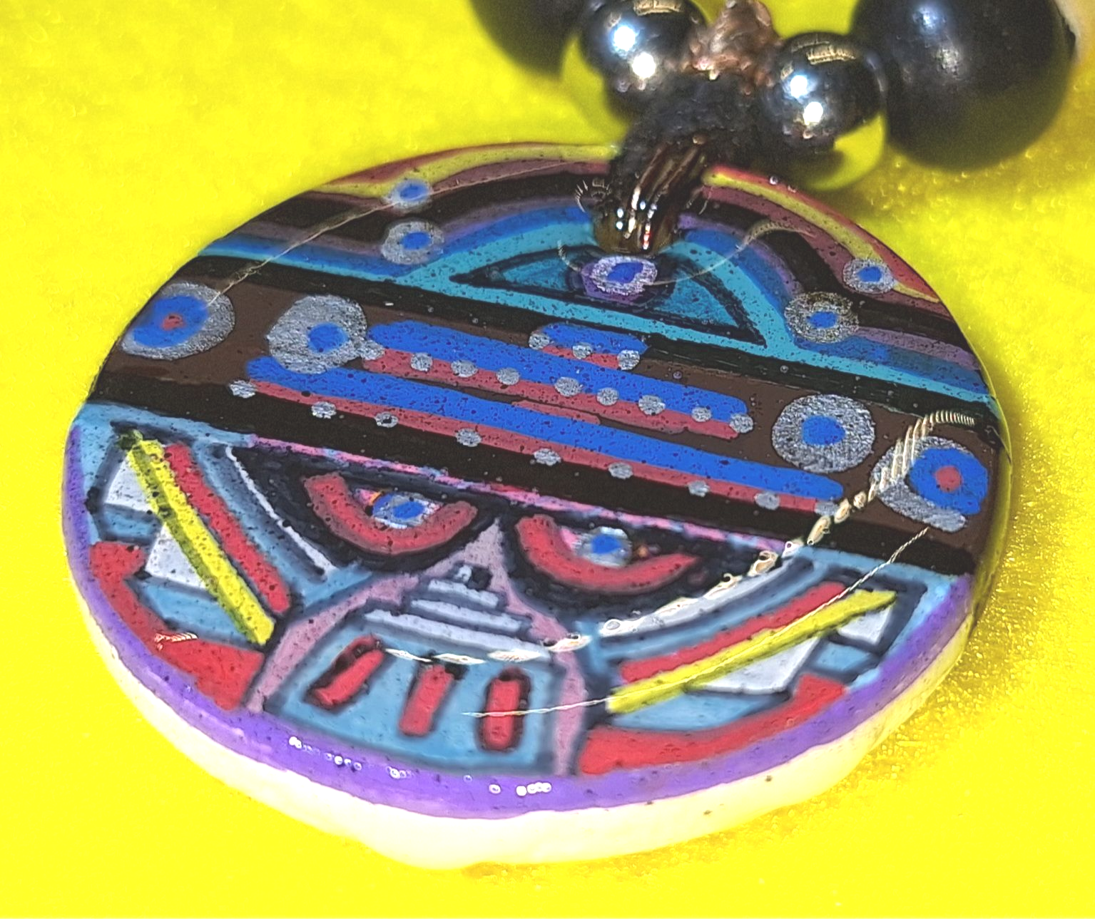 Colorful, patterned pendant with an abstract design, on a beaded necklace, against a yellow backdrop.