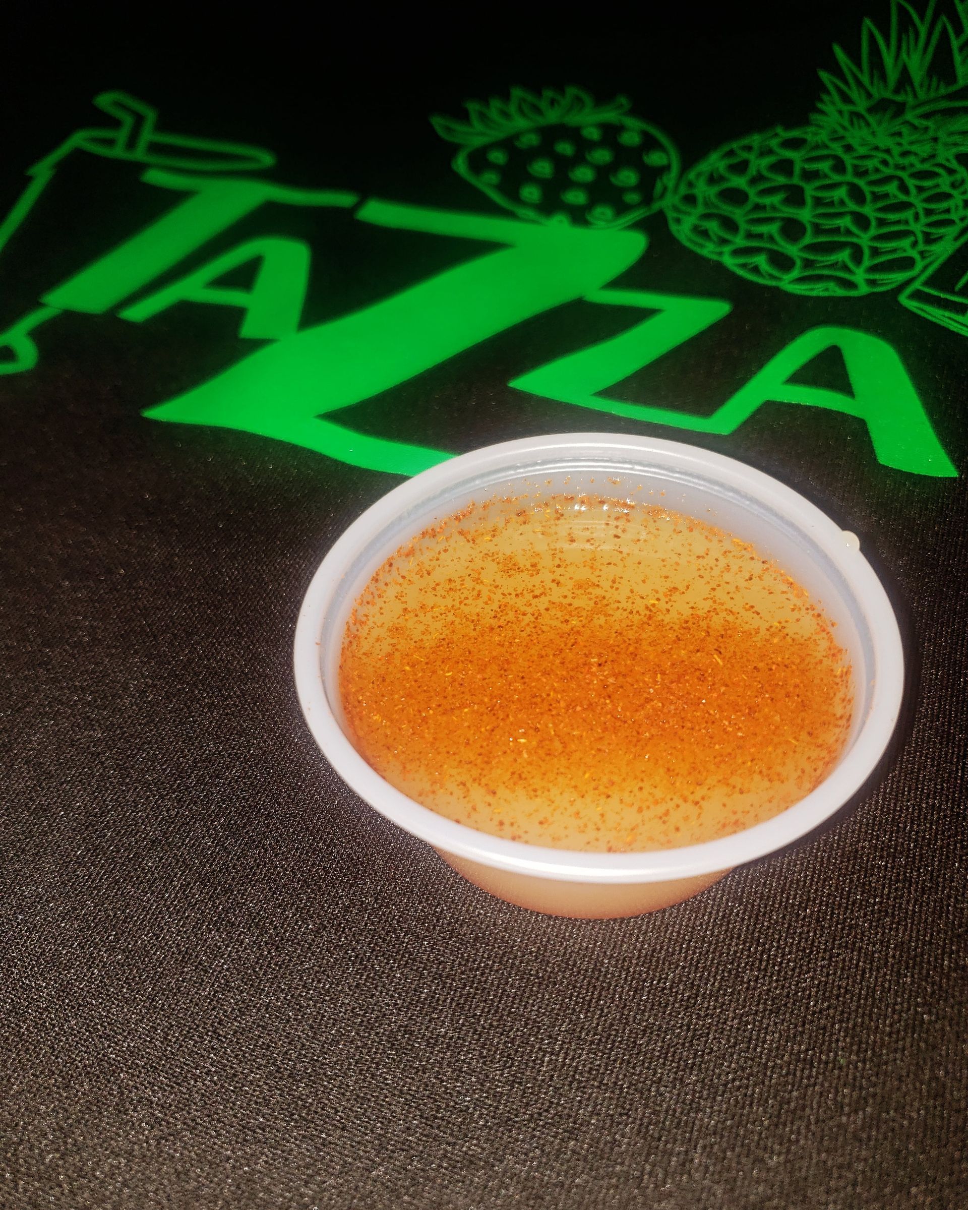 A small cup of sauce sits on a table in front of a sign that says tazza