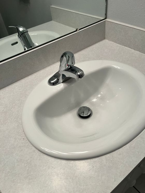 A white bathroom sink and chrome faucet set in a light gray countertop, seen reflected in a mirror.