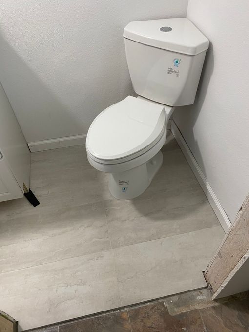 A white corner toilet installed in a small bathroom with light gray flooring and textured walls.