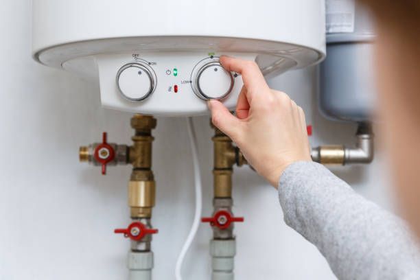 Hand adjusting a white water heater’s control knob above exposed pipes and valves