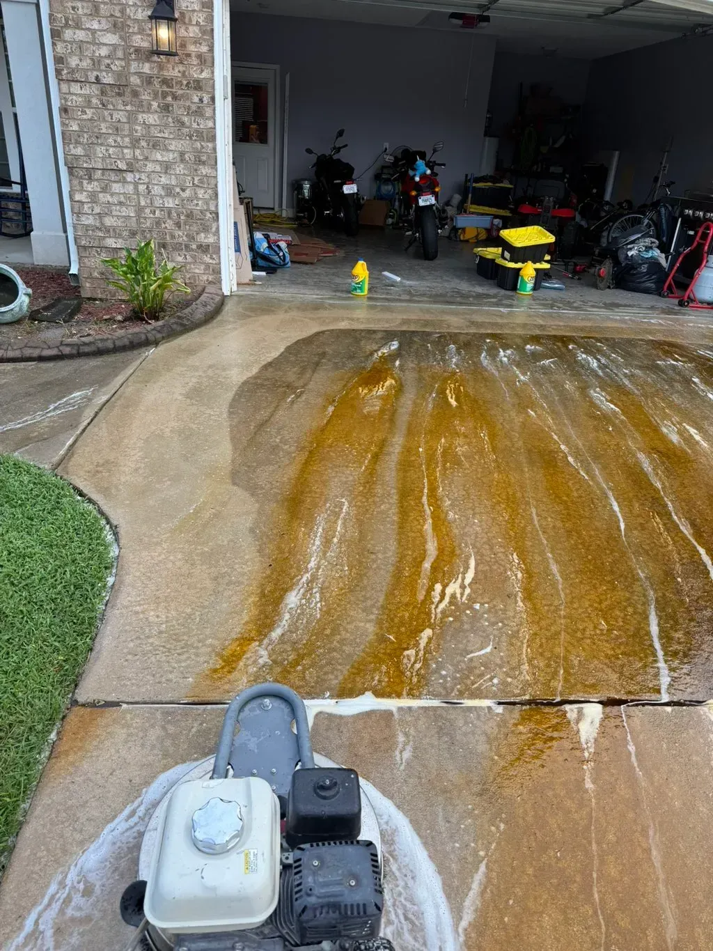 Pressure washing a driveway. Brown and white soapy streaks, green grass, and a garage are visible.
