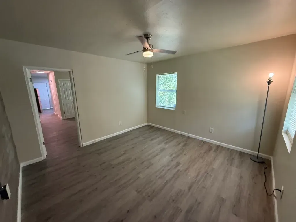 Empty room with gray-toned wood-look flooring, light walls, ceiling fan, and window.