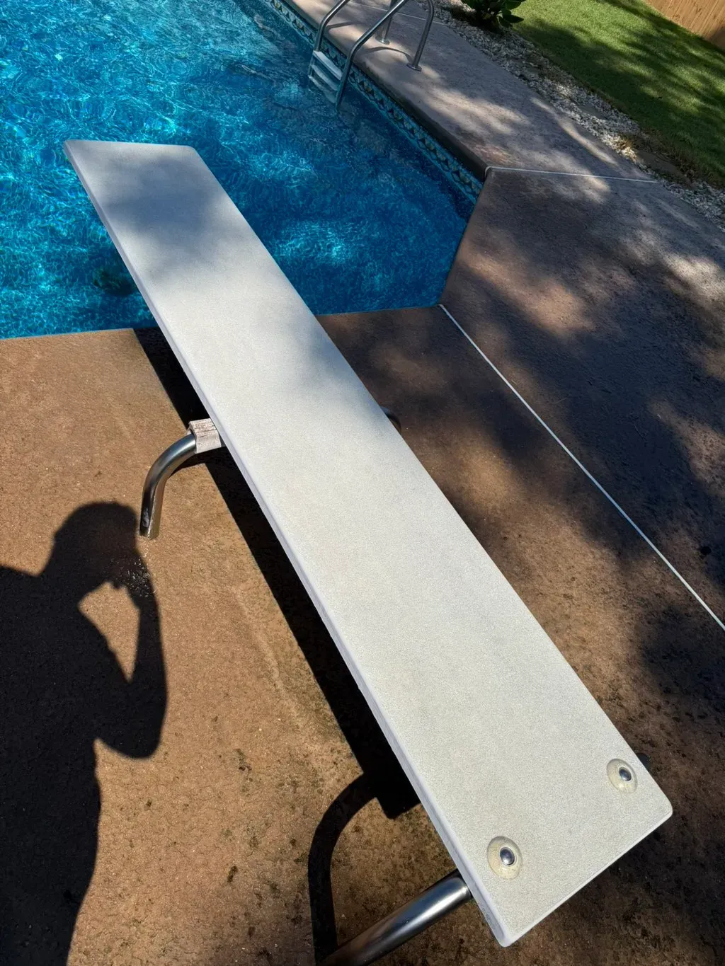 Diving board over a blue pool. The board is light gray, supported by metal. Shadow of a person is visible.