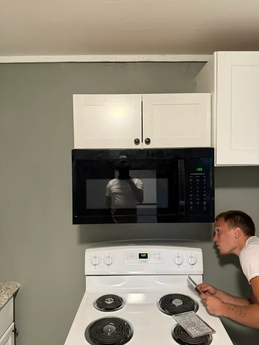 Man working on a stove top below a microwave oven and white kitchen cabinets on a gray wall.