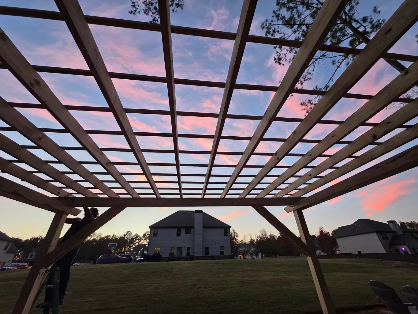 Wooden pergola frame under construction against a colorful sunset sky; person working.