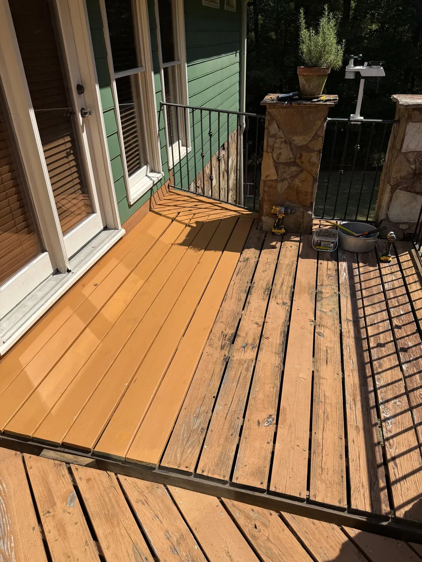 Wooden deck, half-treated, with weathered and freshly coated sections, railing, and adjacent green house.
