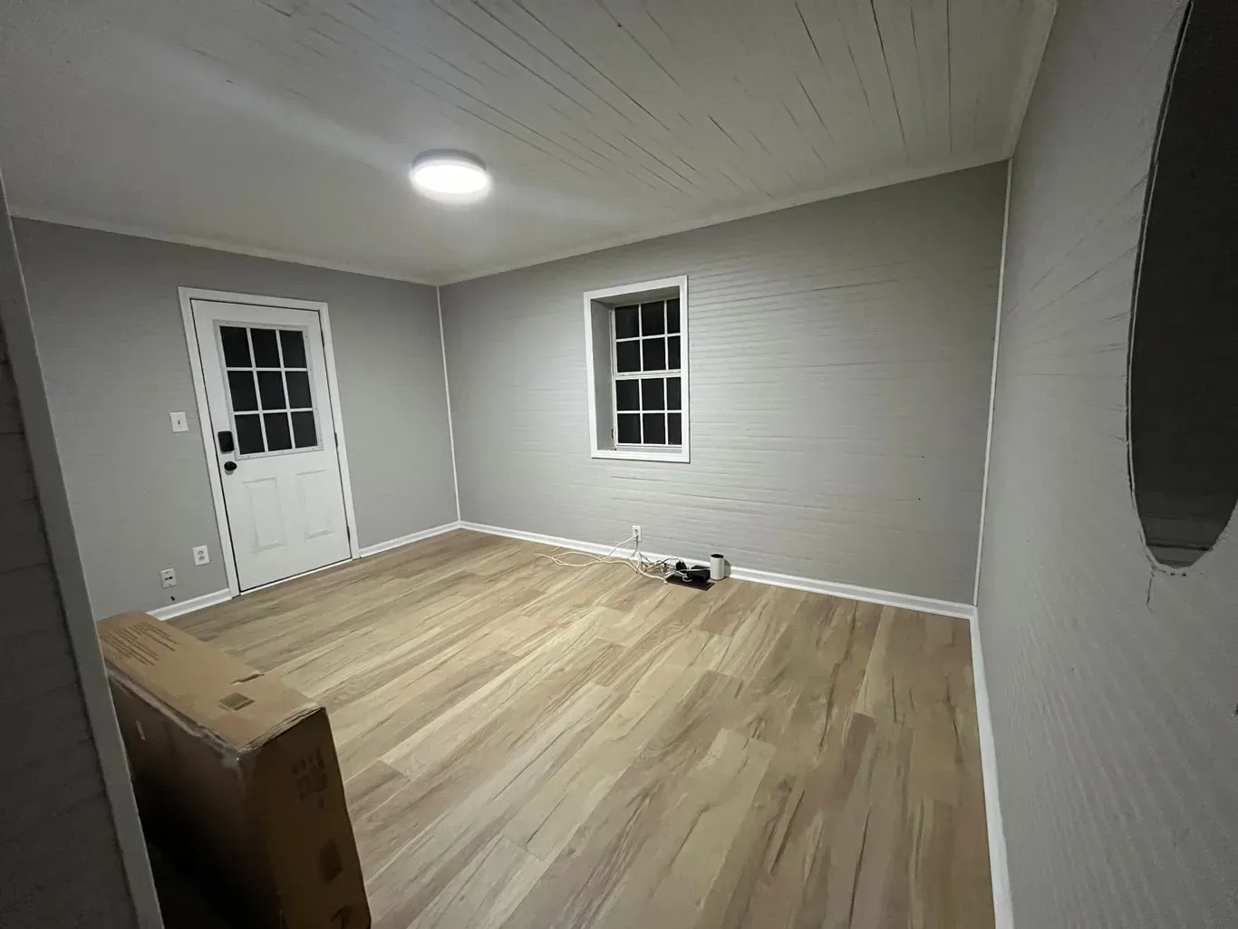 Empty room with light wood floor, gray walls, white door and window, and a circular ceiling light.