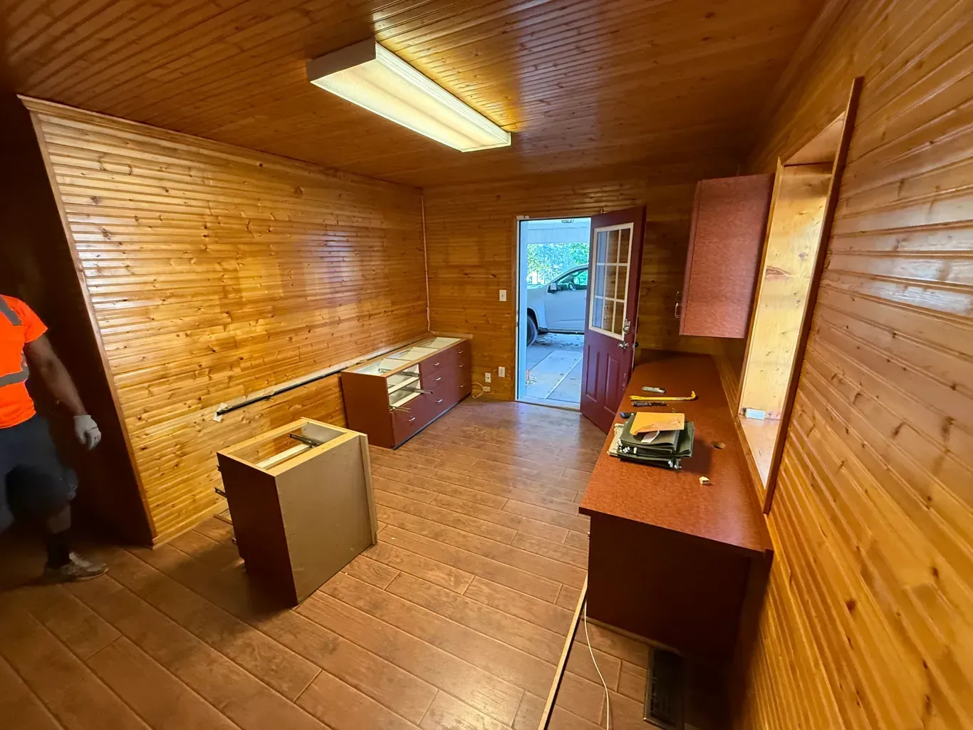 Interior of a wooden paneled room, partially furnished with desks and cabinets. A person in a safety vest is visible.