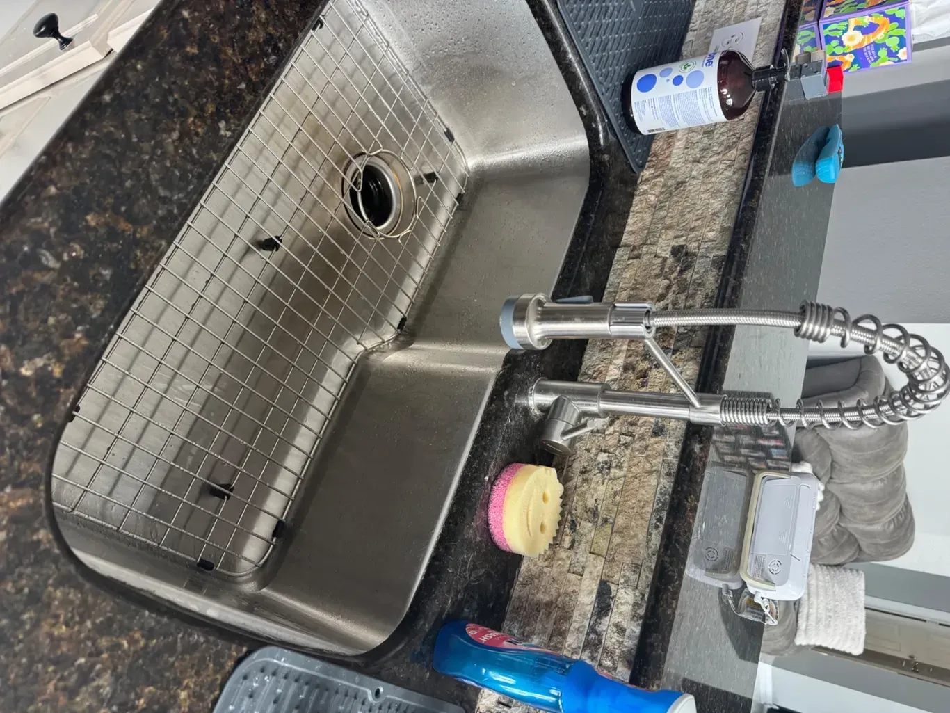 Stainless steel kitchen sink with faucet, dish soap, sponge, and drain strainer on a granite countertop.