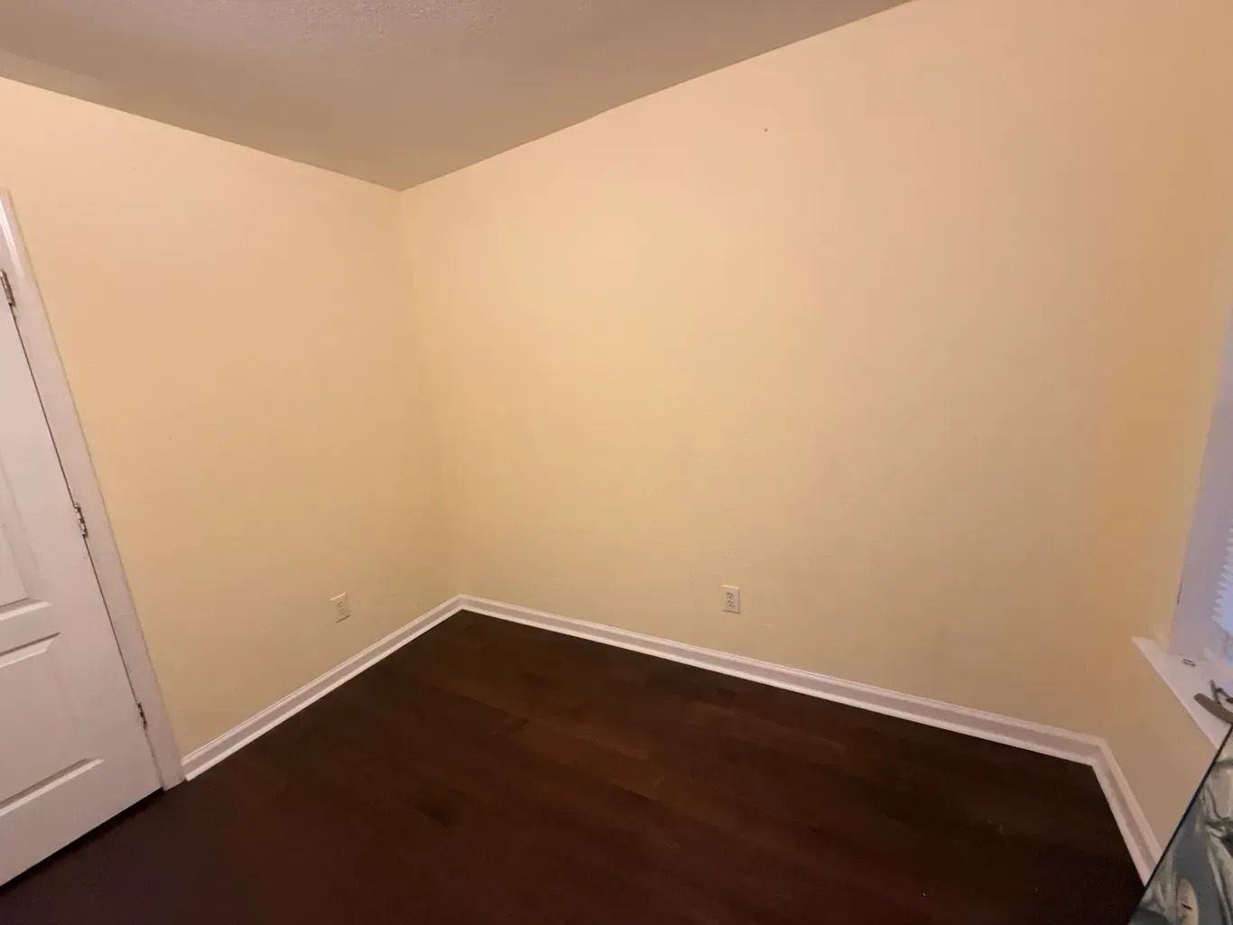 Empty room with tan walls, dark wood floor, white trim, and a closed white door.