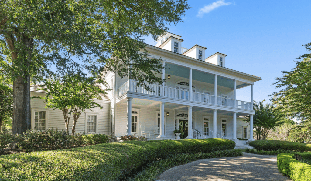 White two-story house with balcony, green bushes in front, tree on the left, clear sky.