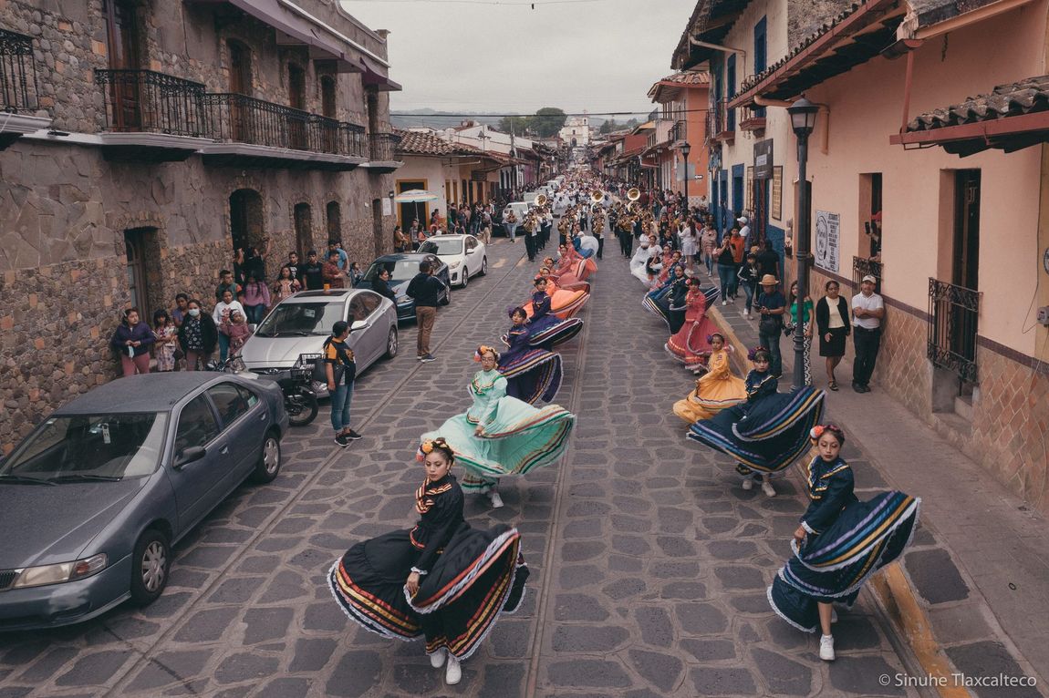 Desfile de bailarines con coloridos trajes tradicionales en una calle empedrada entre edificios.