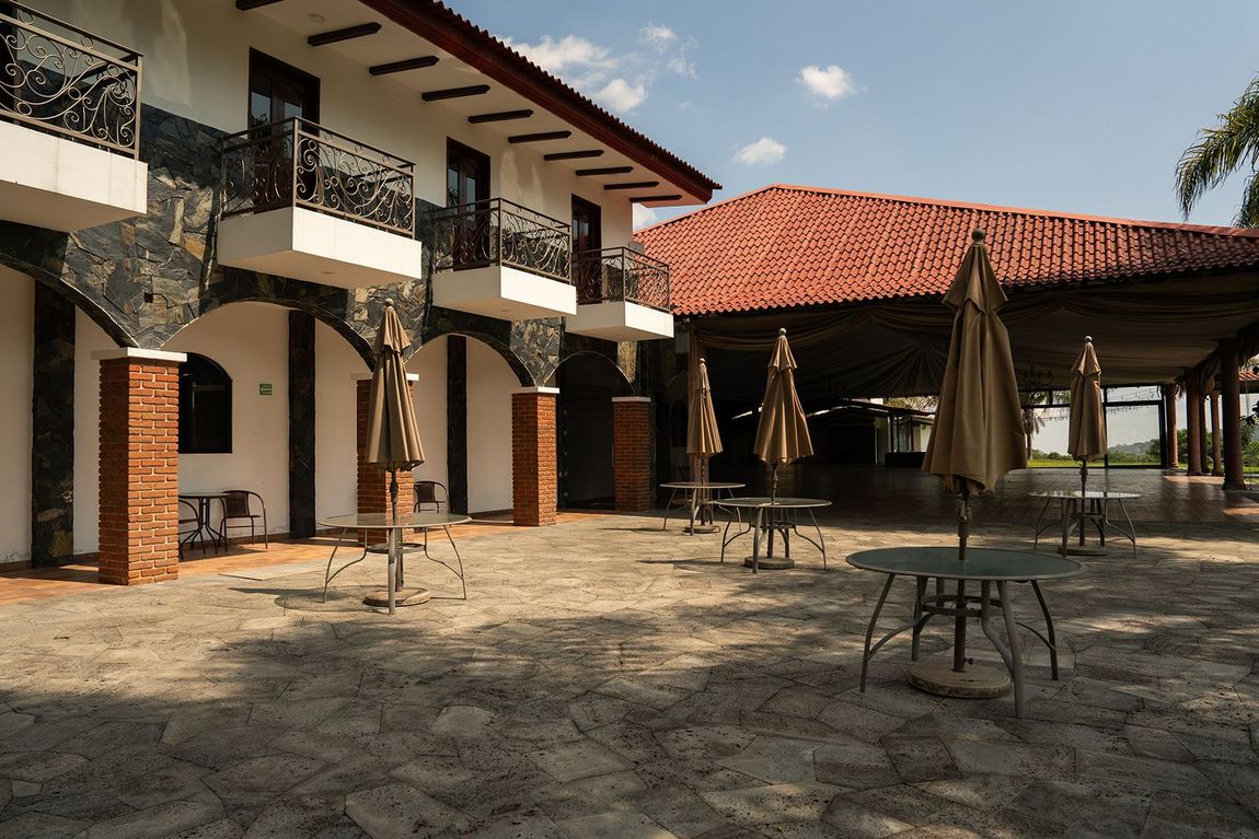 Patio soleado con un edificio de dos plantas, tejado de tejas rojas, mesas y sombrillas cerradas.