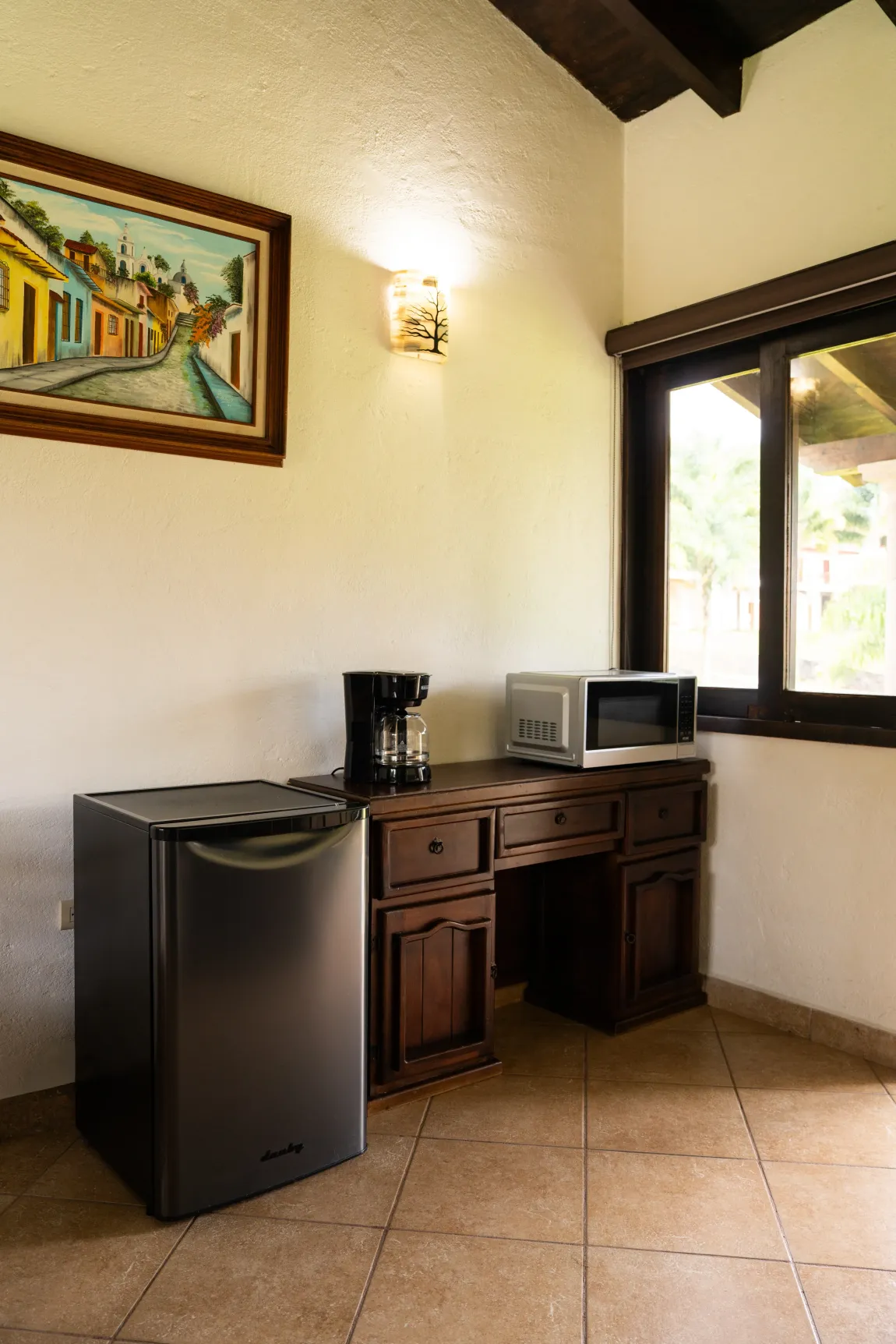 Habitación pequeña con mininevera, cafetera y microondas junto a una ventana y una obra de arte en la pared.