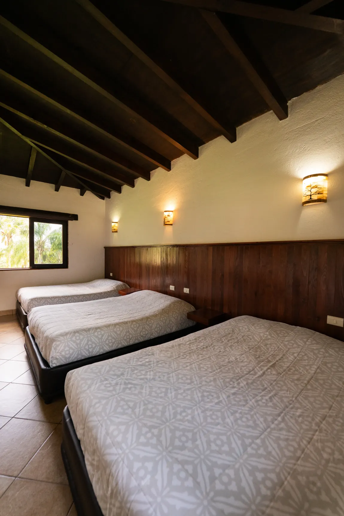 Dormitorio rústico con tres camas bien hechas, paredes revestidas de madera, techo inclinado y apliques de pared.
