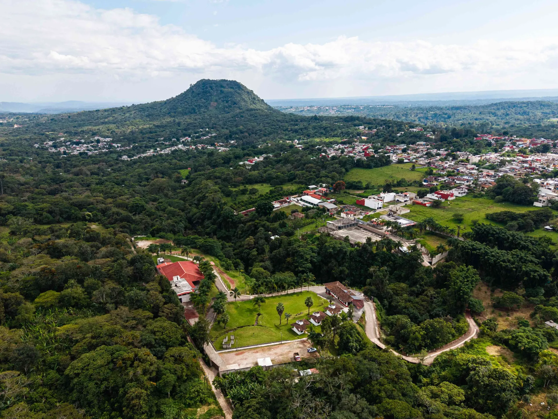Vista aérea de un paisaje verde exuberante con una colina boscosa y casas dispersas bajo un cielo nublado.