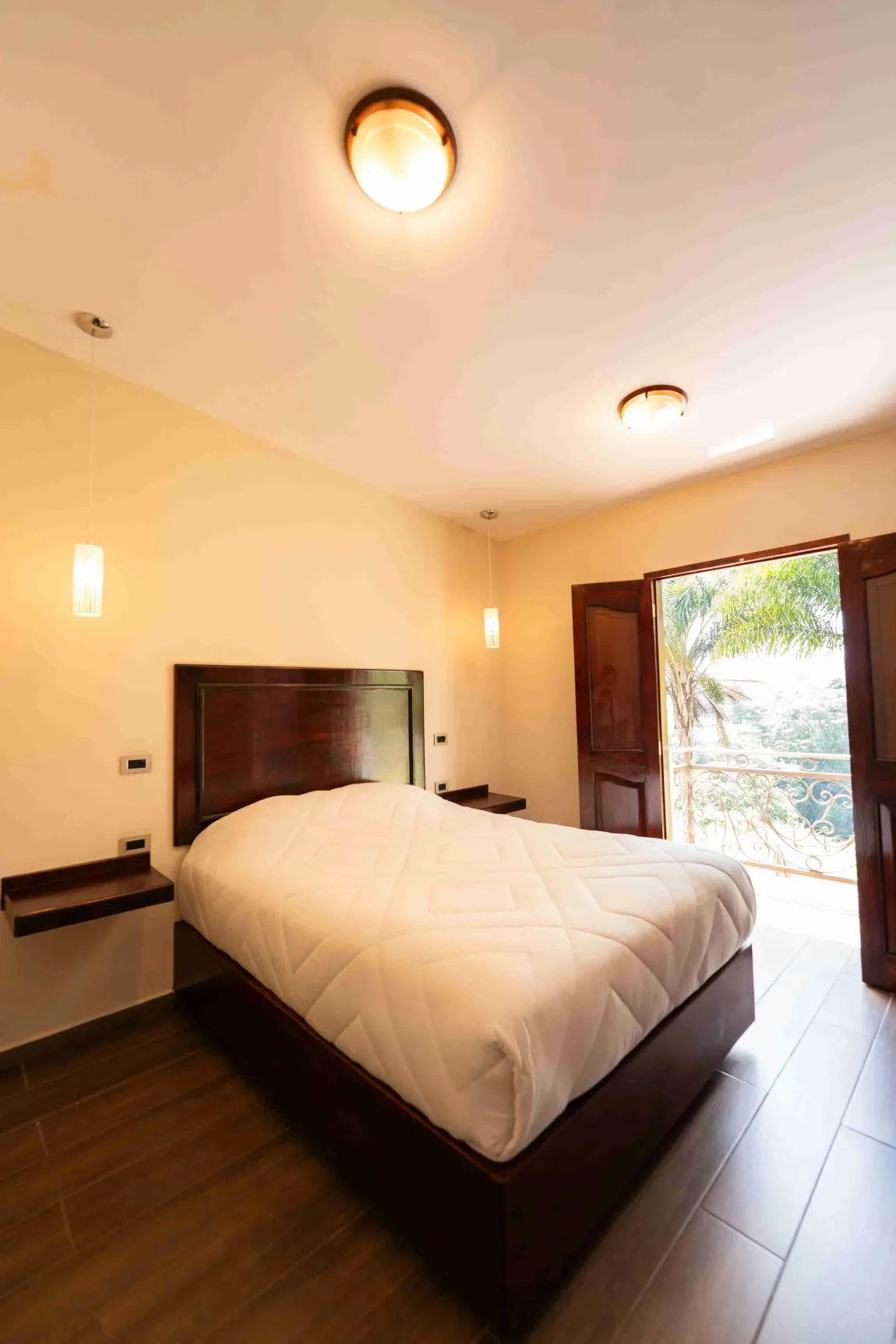 Habitación luminosa con cama, suelo de baldosas y puerta abierta que da a un balcón soleado.