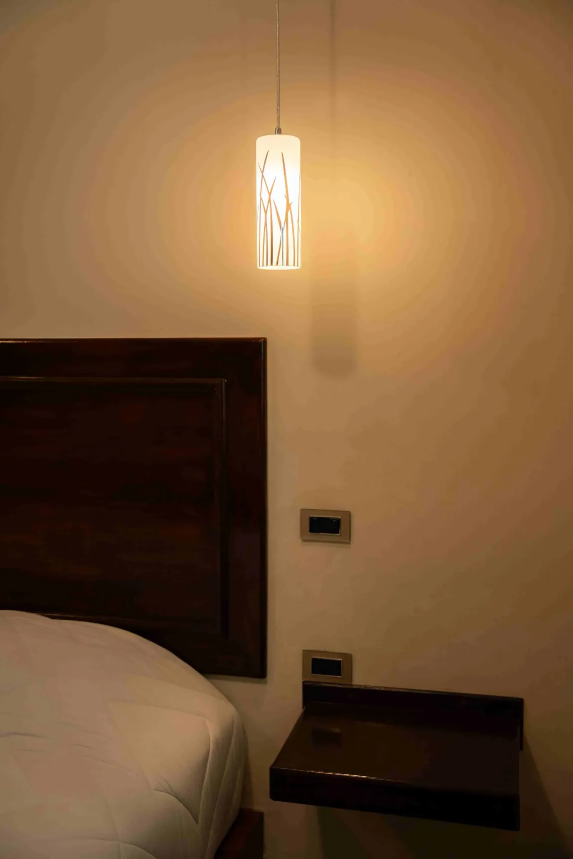 Acogedor rincón de dormitorio con aplique de pared iluminado, cabecero de madera oscura y pequeña mesita de noche.