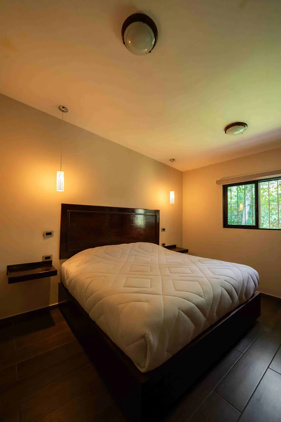 Habitación acogedora con cama grande, cabecero de madera oscura, apliques de pared y ventana con vistas a la vegetación.