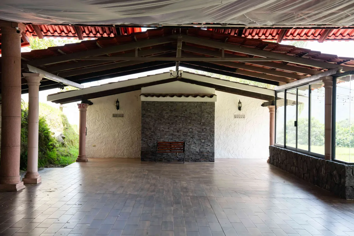 Patio exterior cubierto con suelo de baldosas, vigas de madera y muro de piedra en la parte trasera.