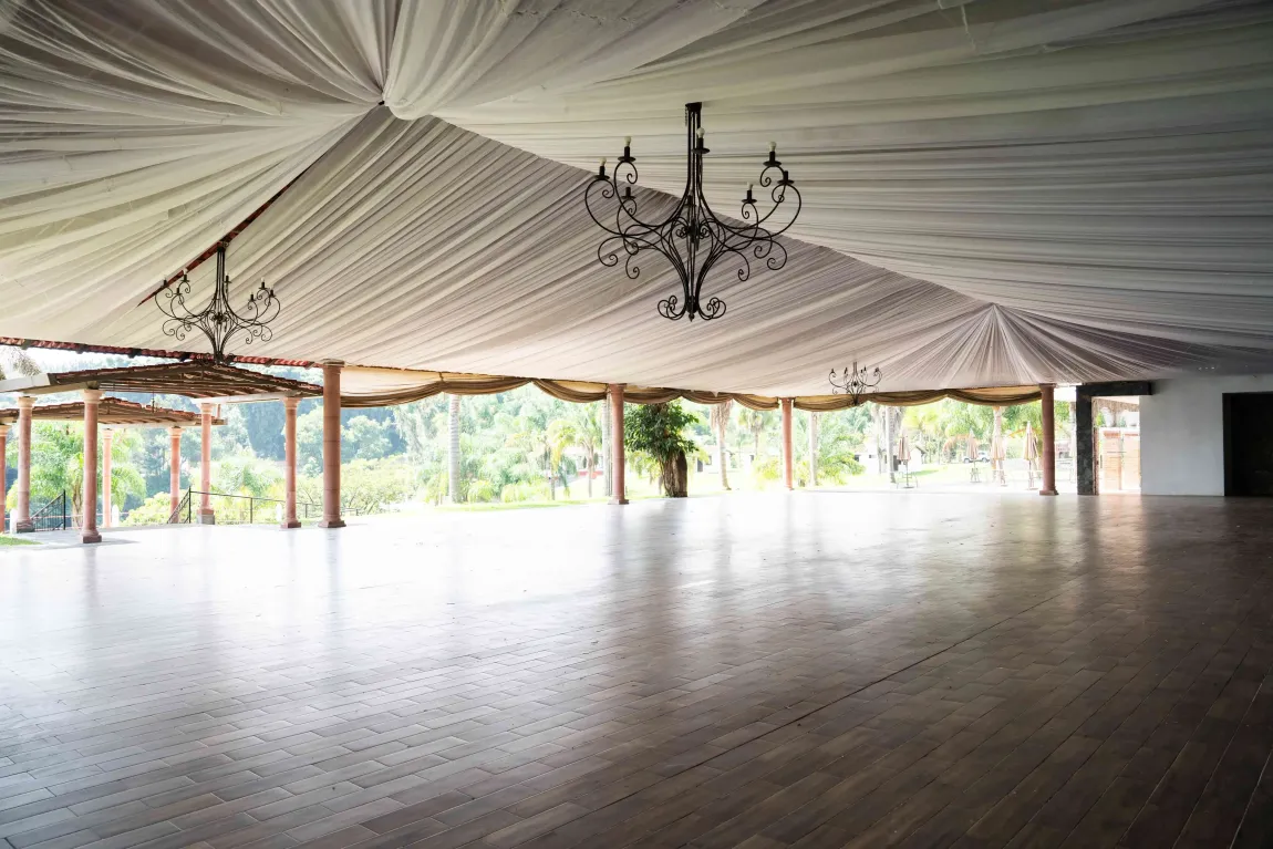 Salón interior vacío con techo cubierto de cortinas blancas, candelabros y grandes ventanales con vistas a zonas verdes.