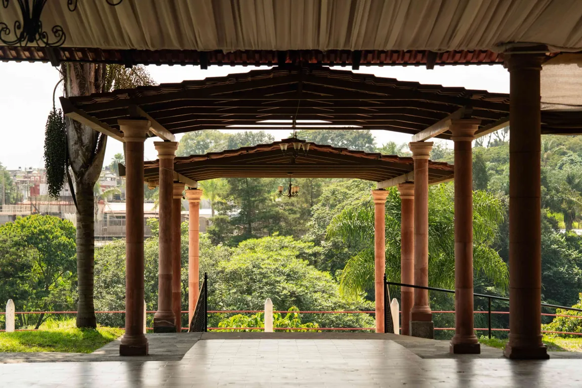 Pabellón al aire libre con columnas de madera y pérgola con vistas a frondosos árboles verdes.