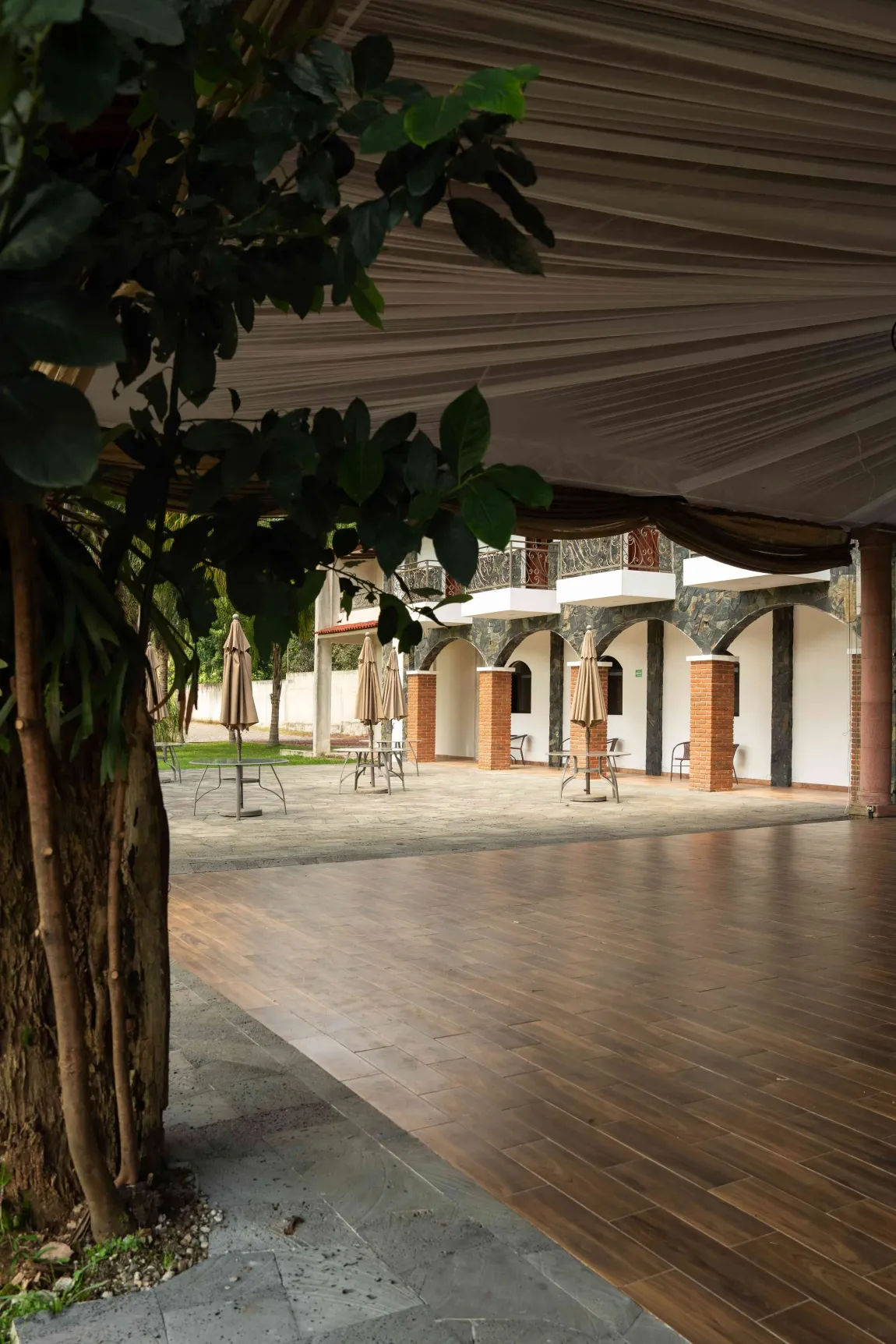 Terraza de madera empapada por la lluvia junto a un edificio blanco, vista desde debajo de un árbol y un toldo.