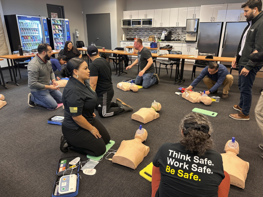 People kneeling in a training room practicing CPR on mannequins, with a “Think Safe Work Safe” vest in the foreground
