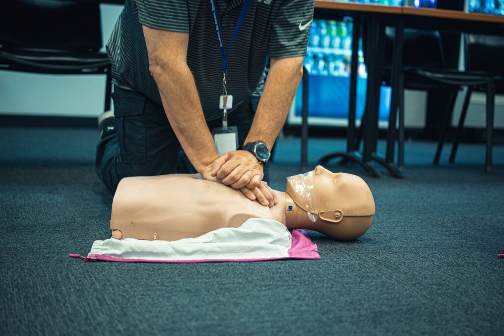 Person kneeling beside a CPR training mannequin on a carpeted floor, demonstrating chest compressions