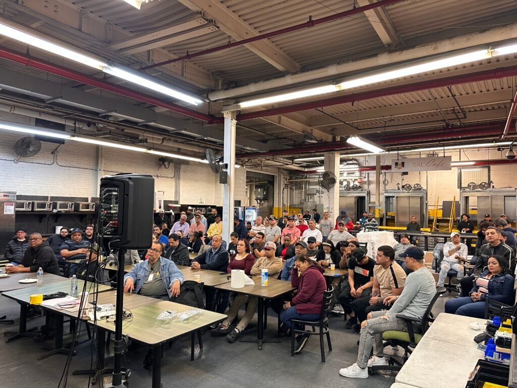 Crowded industrial hall with people seated at long tables, listening to a speaker near the front.