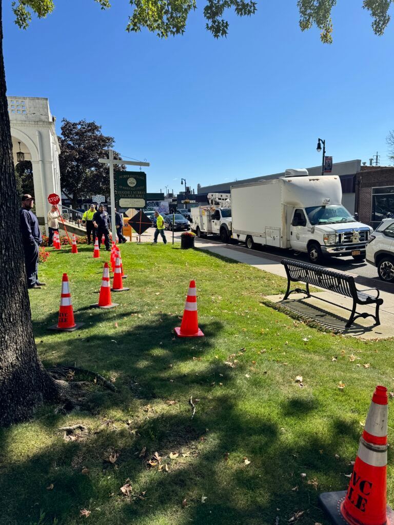 Workers and cones line a sunny sidewalk beside parked trucks and a building.