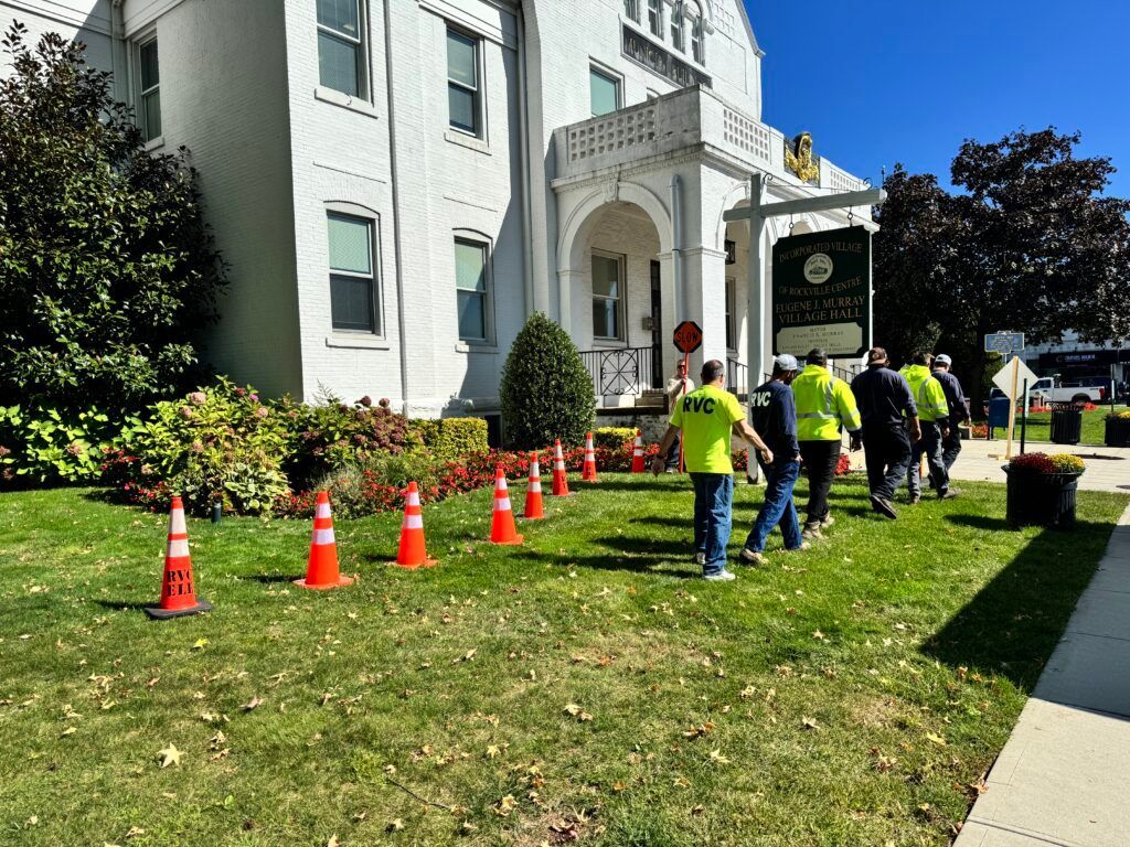 Workers in neon vests set orange cones outside a white building with an arched entrance.