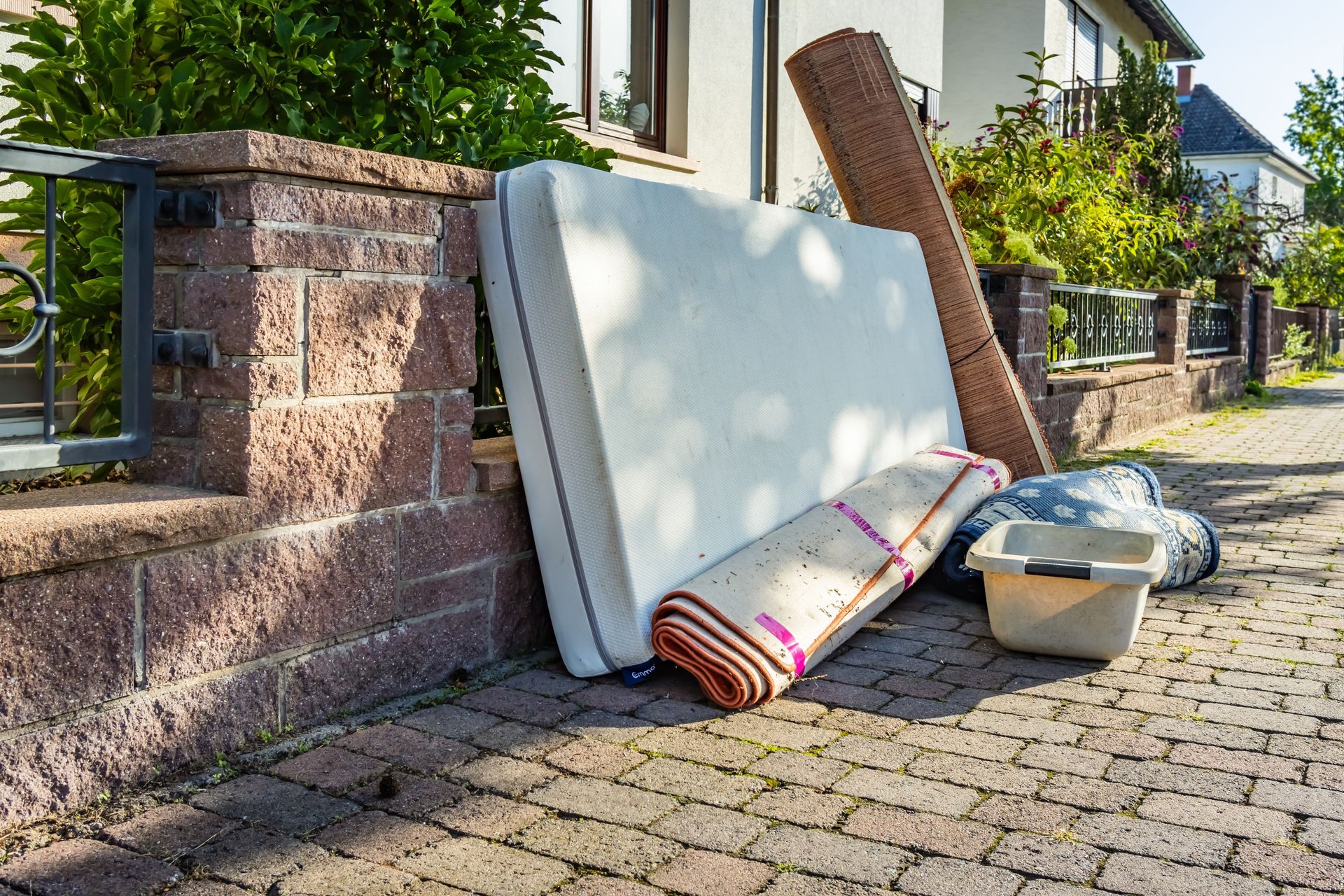 Discarded mattresses and a metal bed frame on a sidewalk beside a brick wall and house fence.