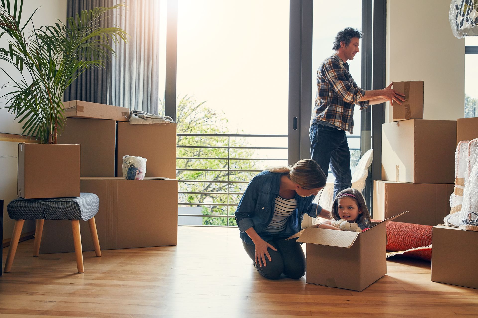 Family unpacking moving boxes in a sunlit living room, with a child sitting in one box.