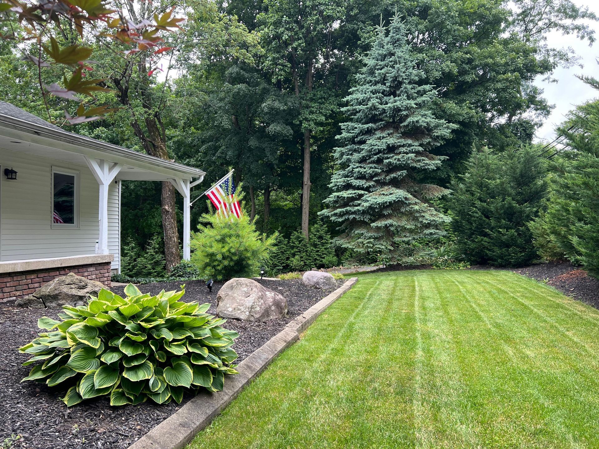 Landscaped front yard with manicured lawn, hostas, and trees beside a house porch