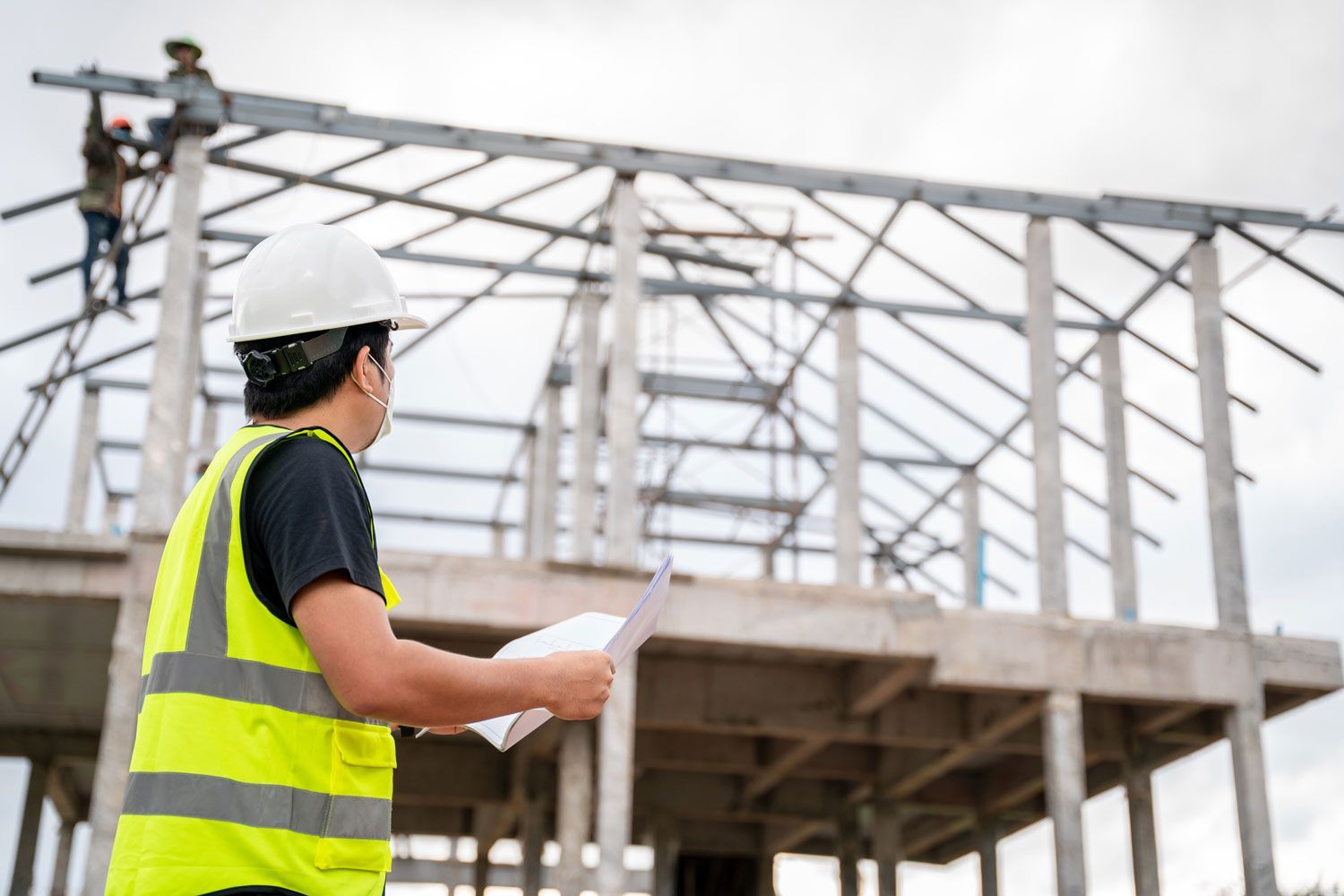 Construction worker in yellow vest and hard hat reviewing plans at a building site as workers construct the roof.