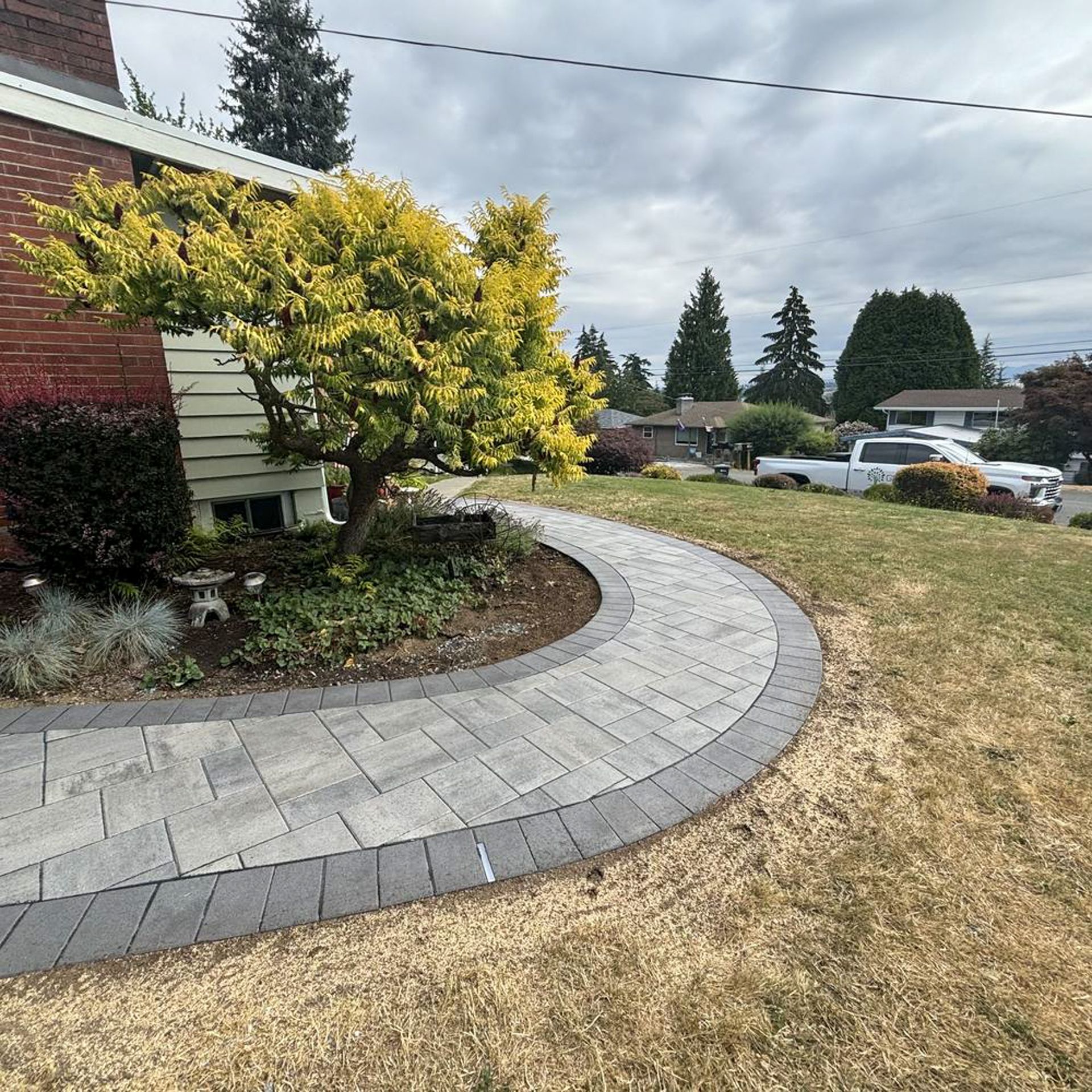 Curving paved walkway leading to a house with a small tree in a landscaped bed. Cloudy sky.