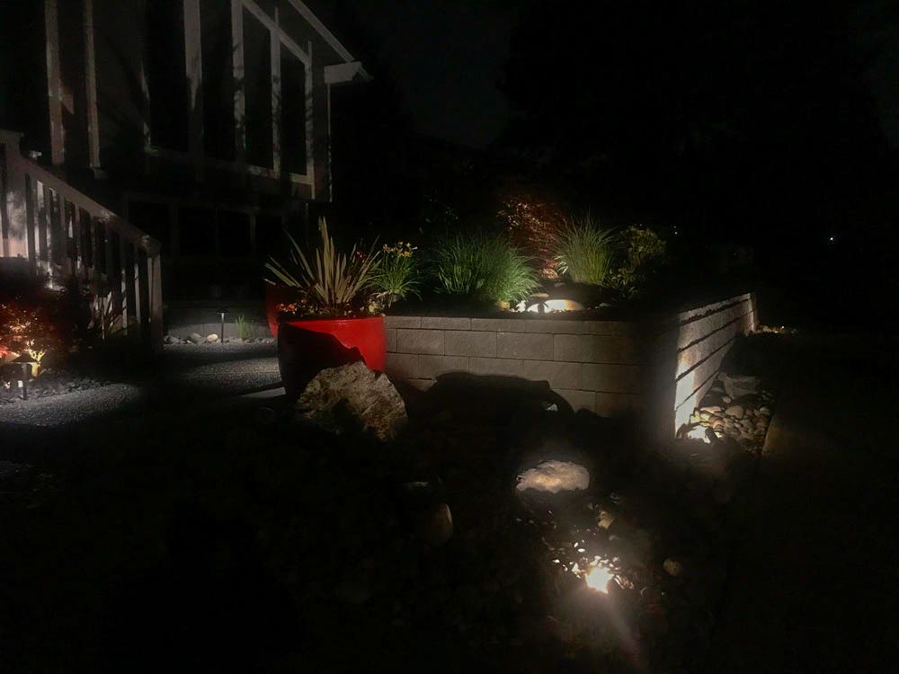 Night view of a house's illuminated landscaping; red pot with plants, stone path, and raised garden bed.