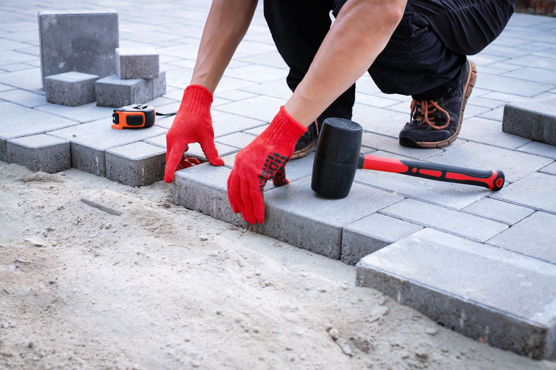 Person in red gloves laying paving stones on sand, tools nearby.