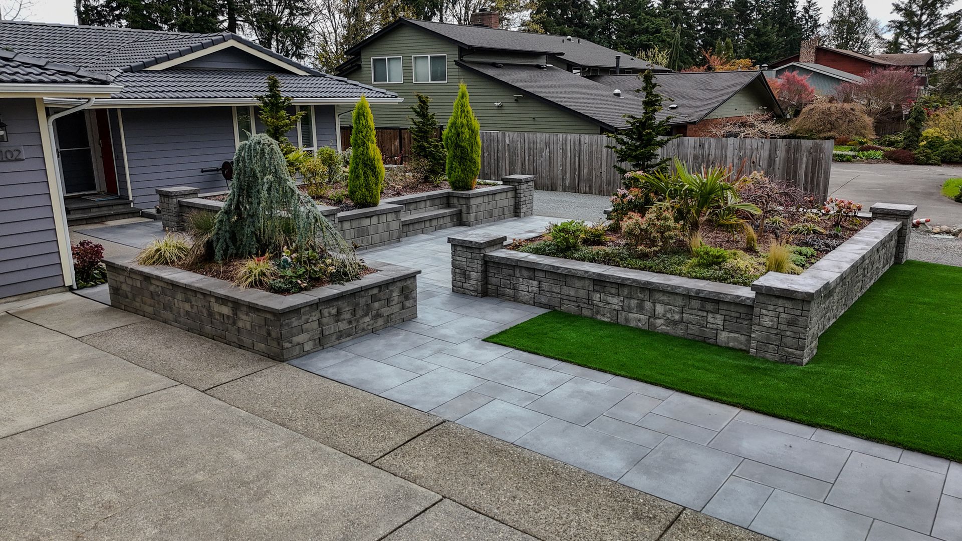 Stone planters with greenery on concrete patio in front of houses.