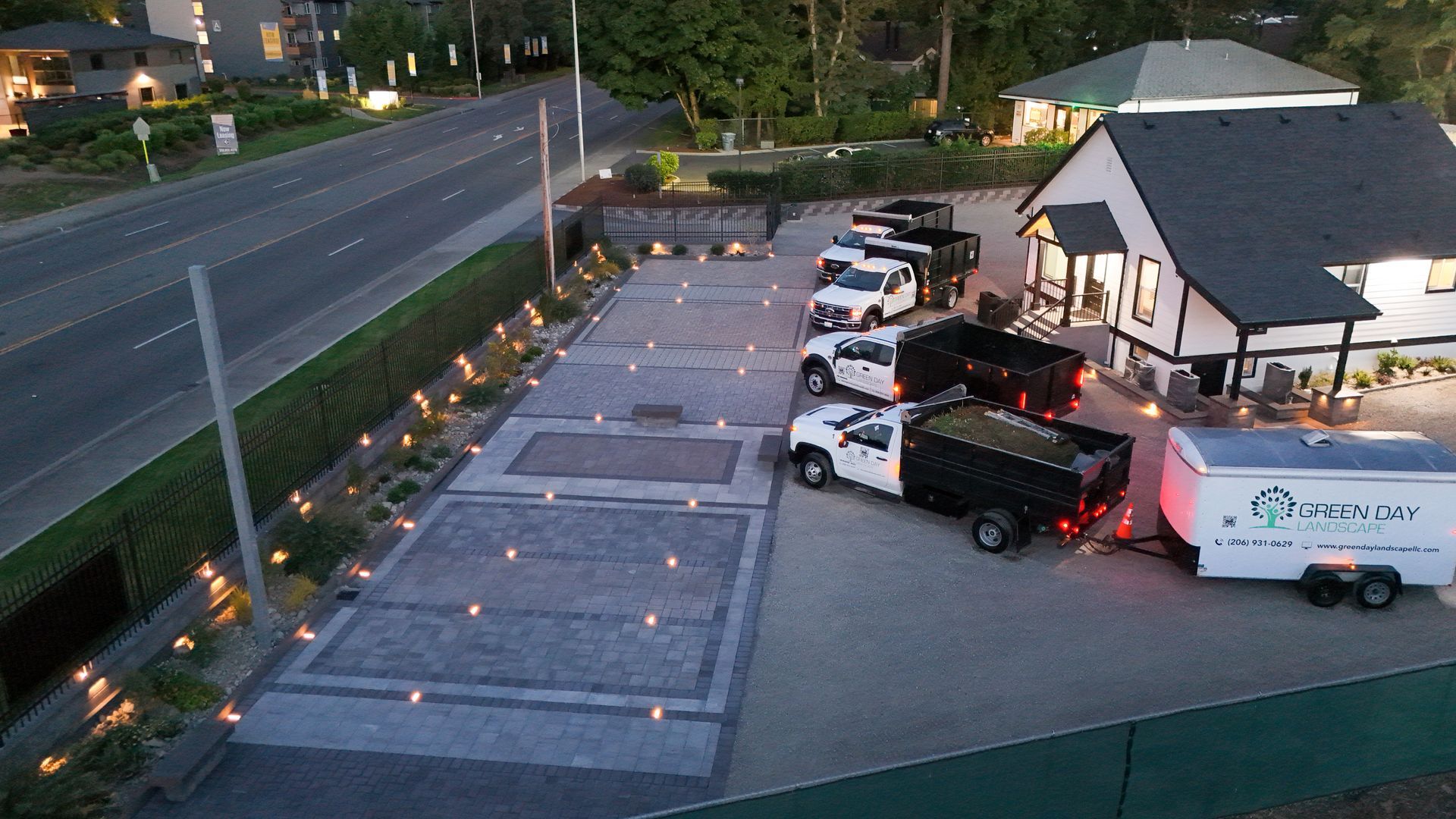 Vehicles at a paved lot next to a building, next to a road. Dump trucks and trailer visible. Dusk.