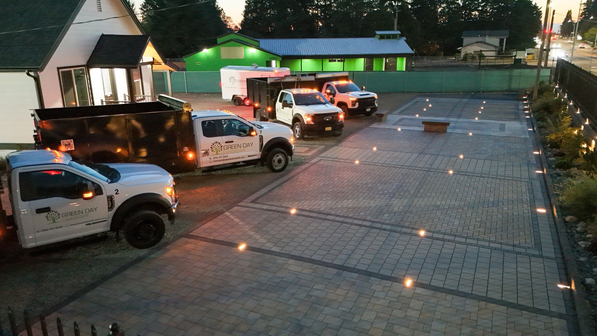 Four white trucks parked on a brick paved lot at dusk, near a building and greenery, with small lights in the ground.