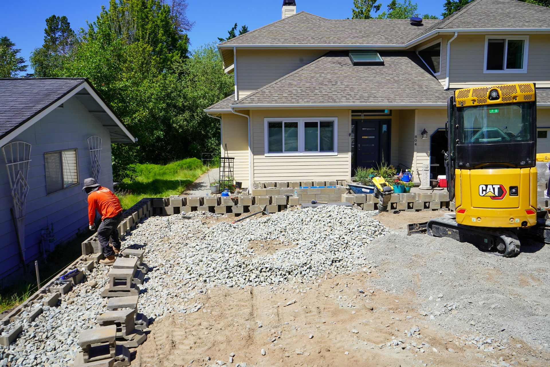 Construction of a driveway with a mini excavator and worker laying blocks near a two-story house.