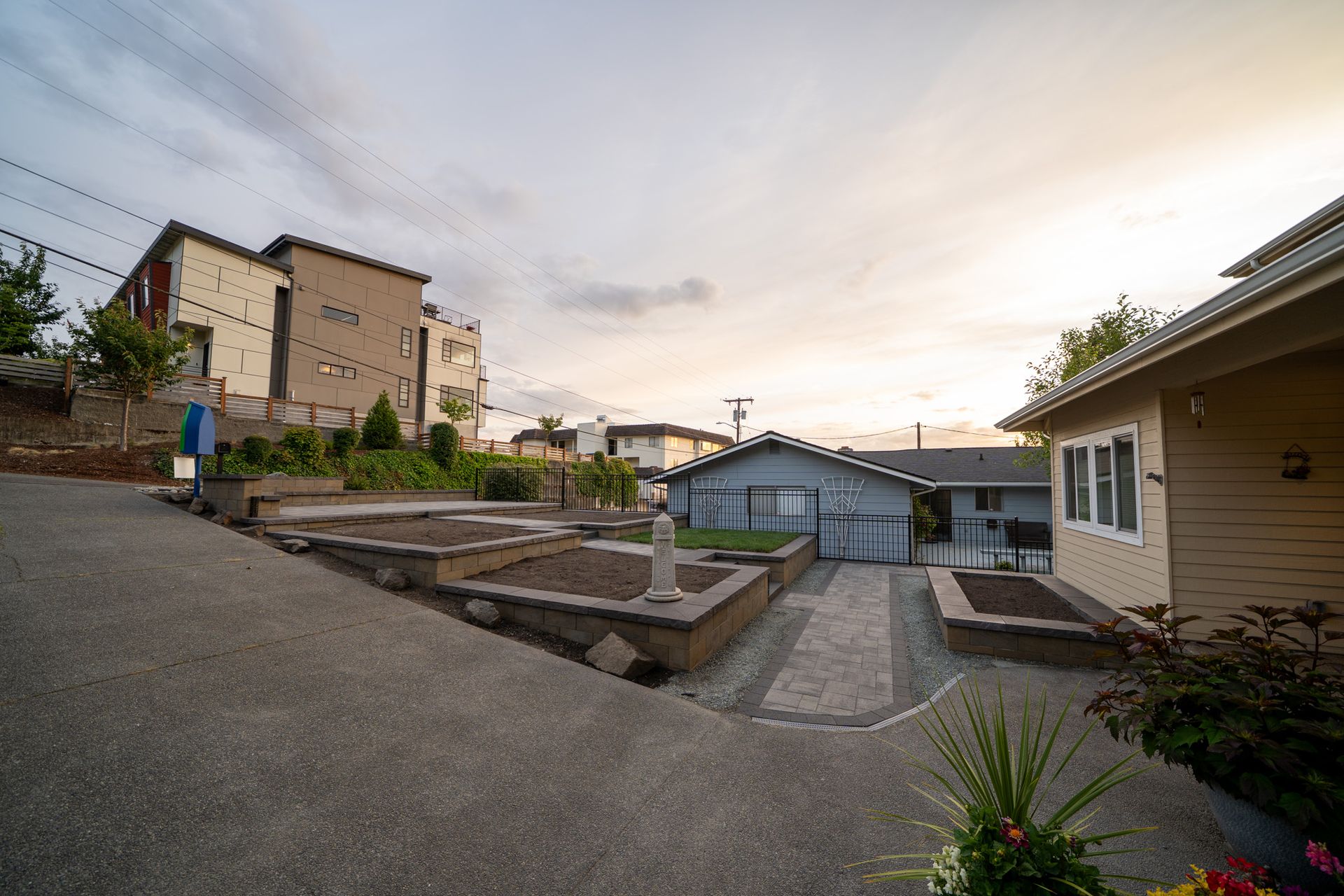 Raised garden beds and buildings on a sloping paved lot under a cloudy sky.