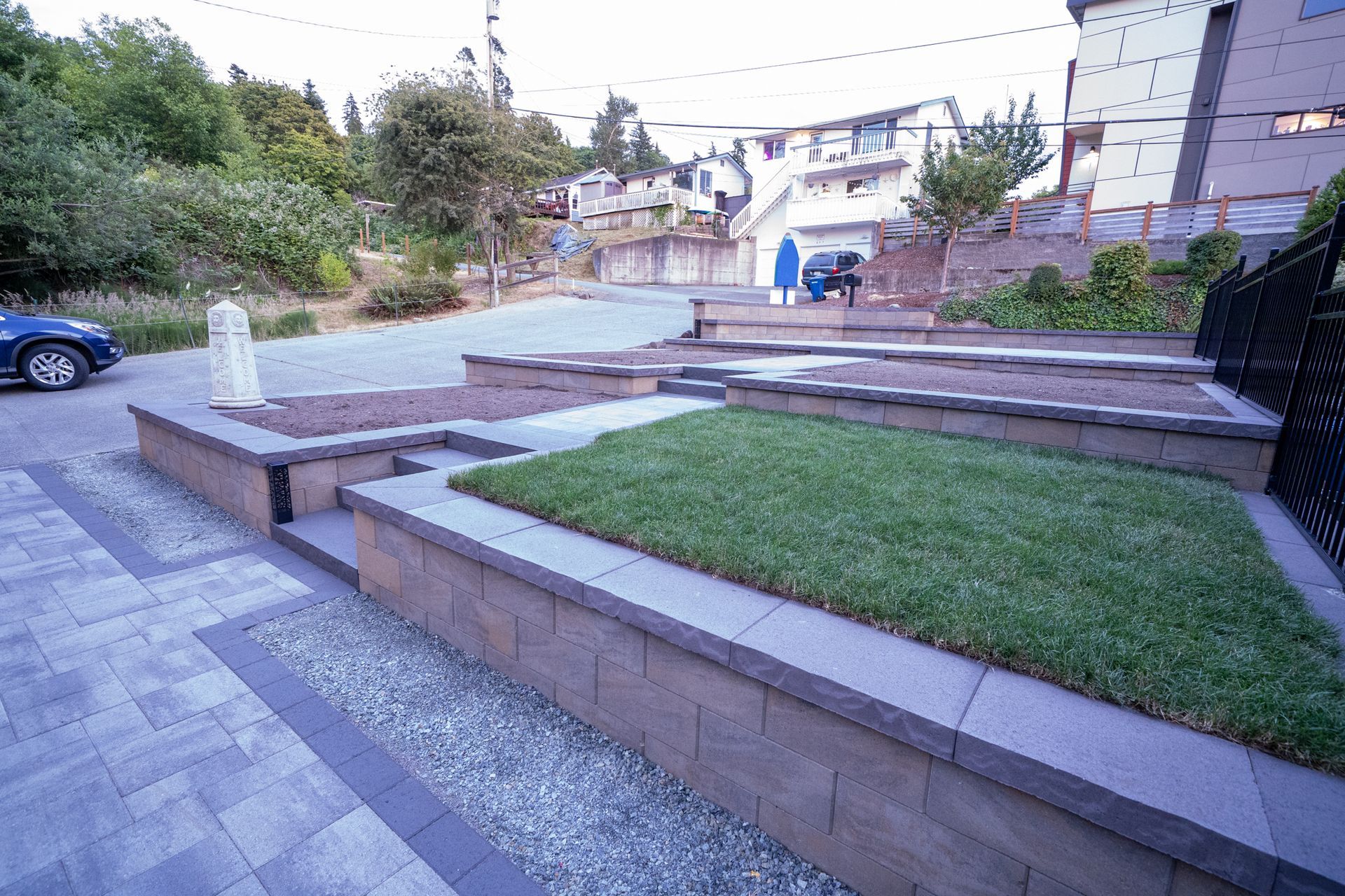 Landscaped front yard with tiered planters, grass, and gravel. Blue car parked on the left.