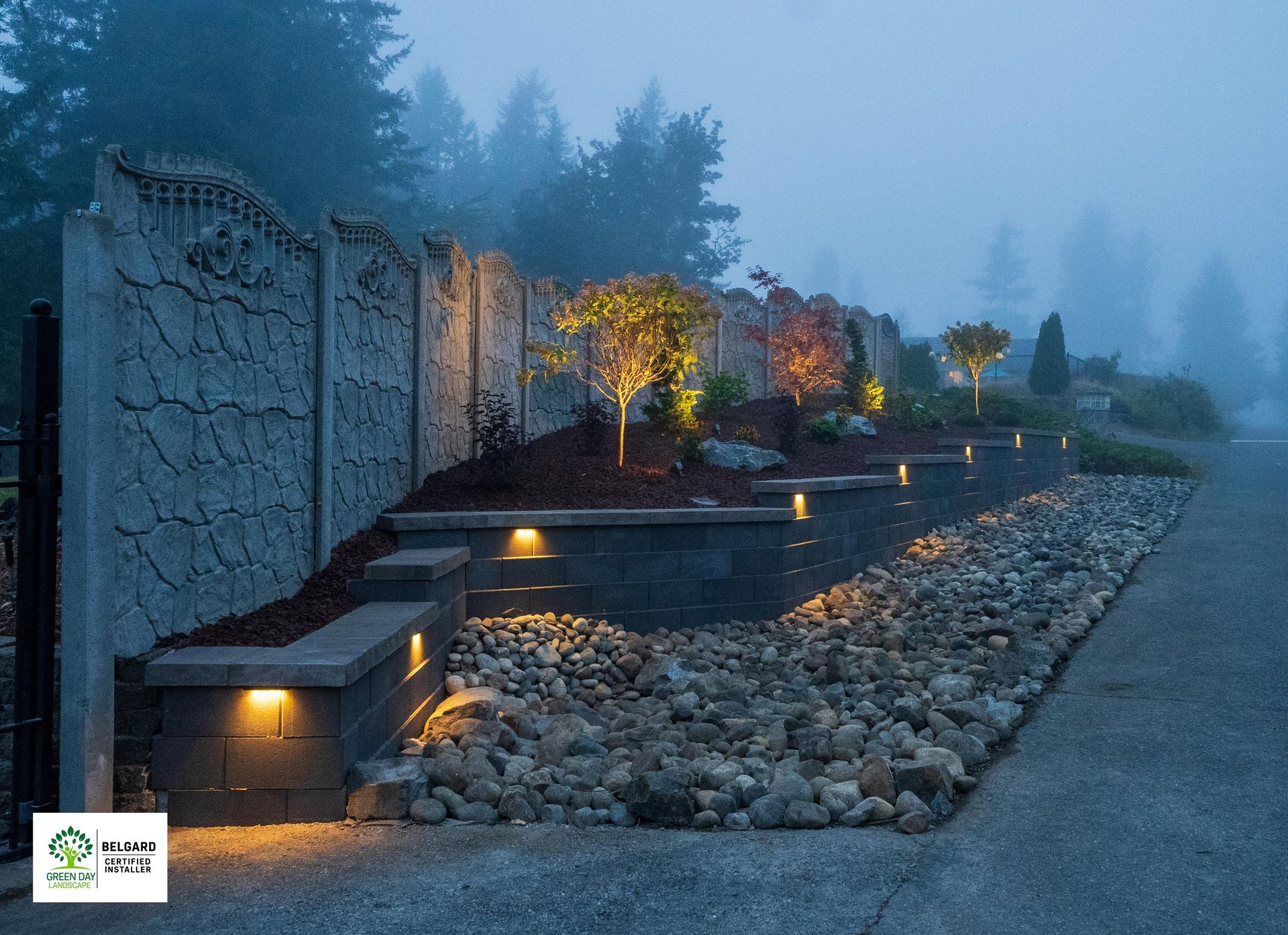 Lit landscape with stone wall, trees, and rock pathway on a foggy evening.