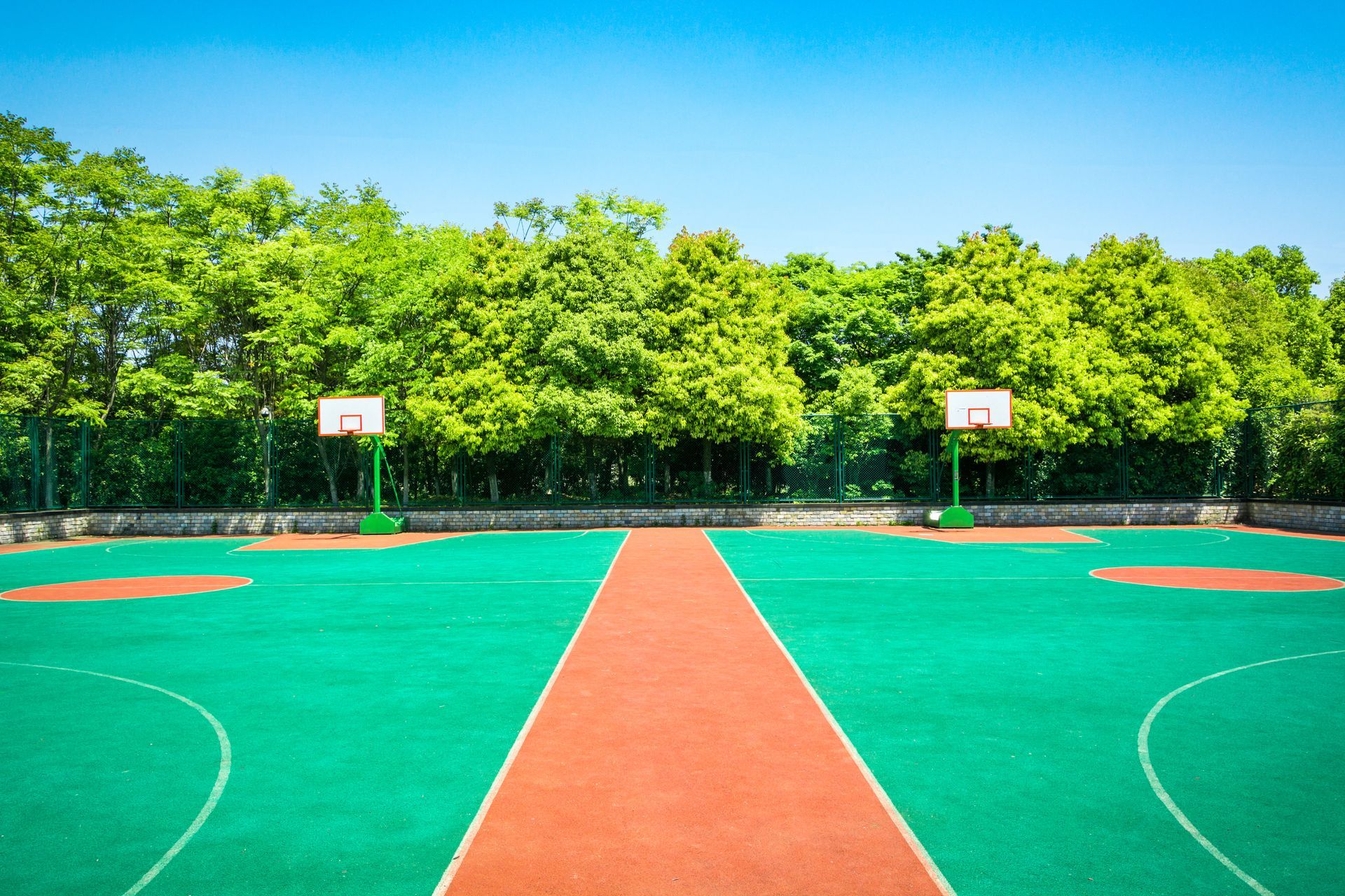 Basketball court with green surface and two hoops; trees in the background under a blue sky.